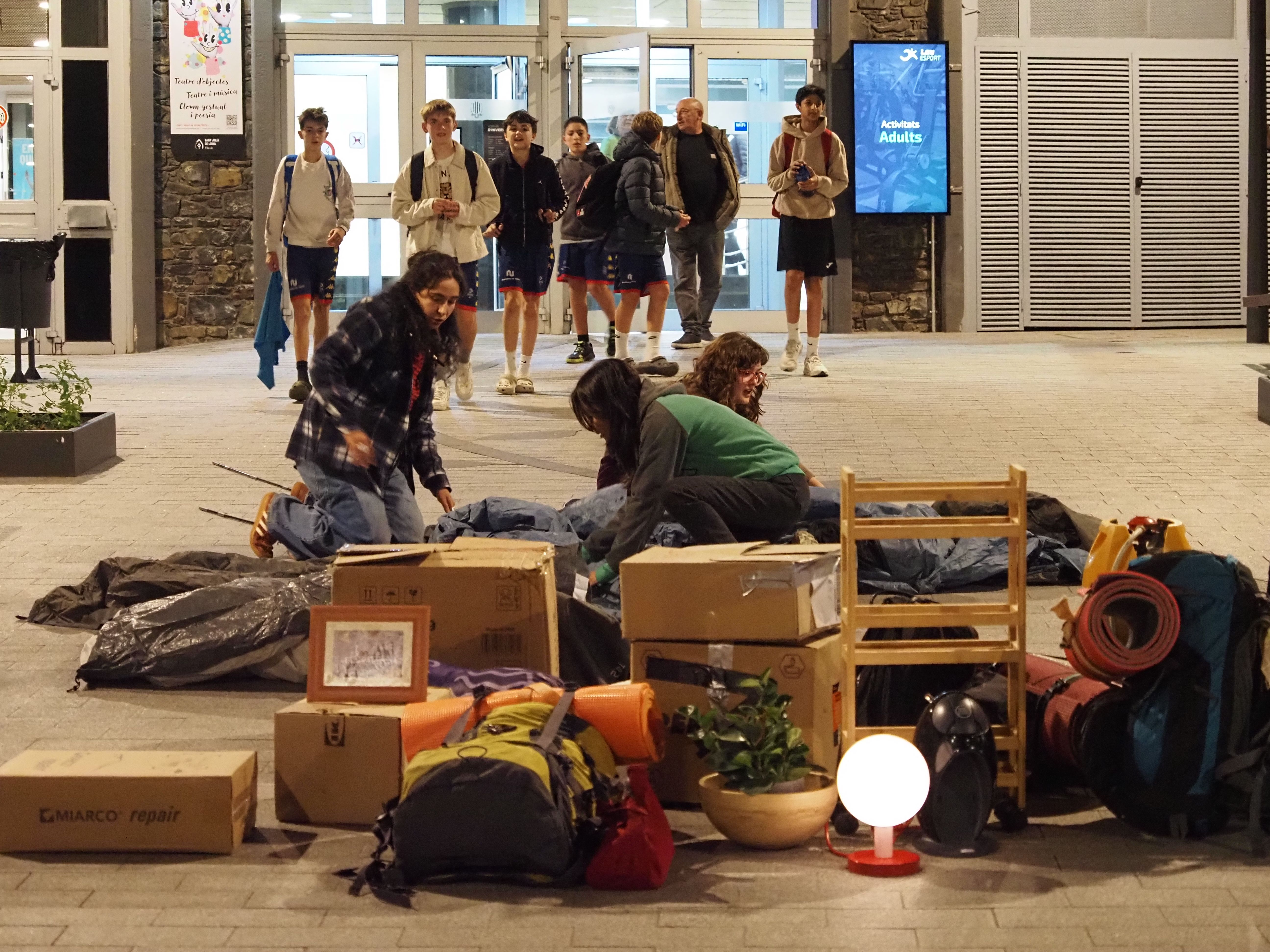 Les tres actrius començant a muntar la tenda amb un grup de joves aliens a tot plegat sortint del centre esportiu.