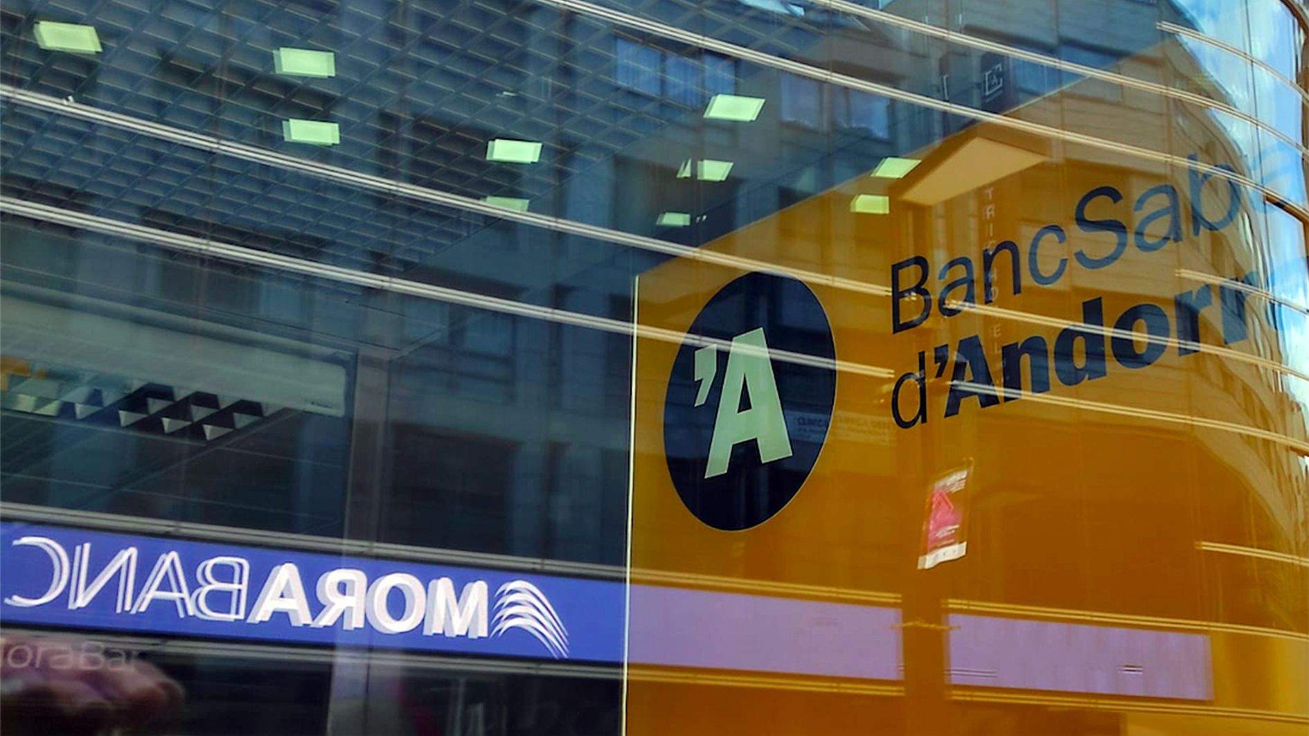 Els rètols de BancSabadell d'Andorra i MoraBanc reflectits en la vidriera d'una oficina del primer.